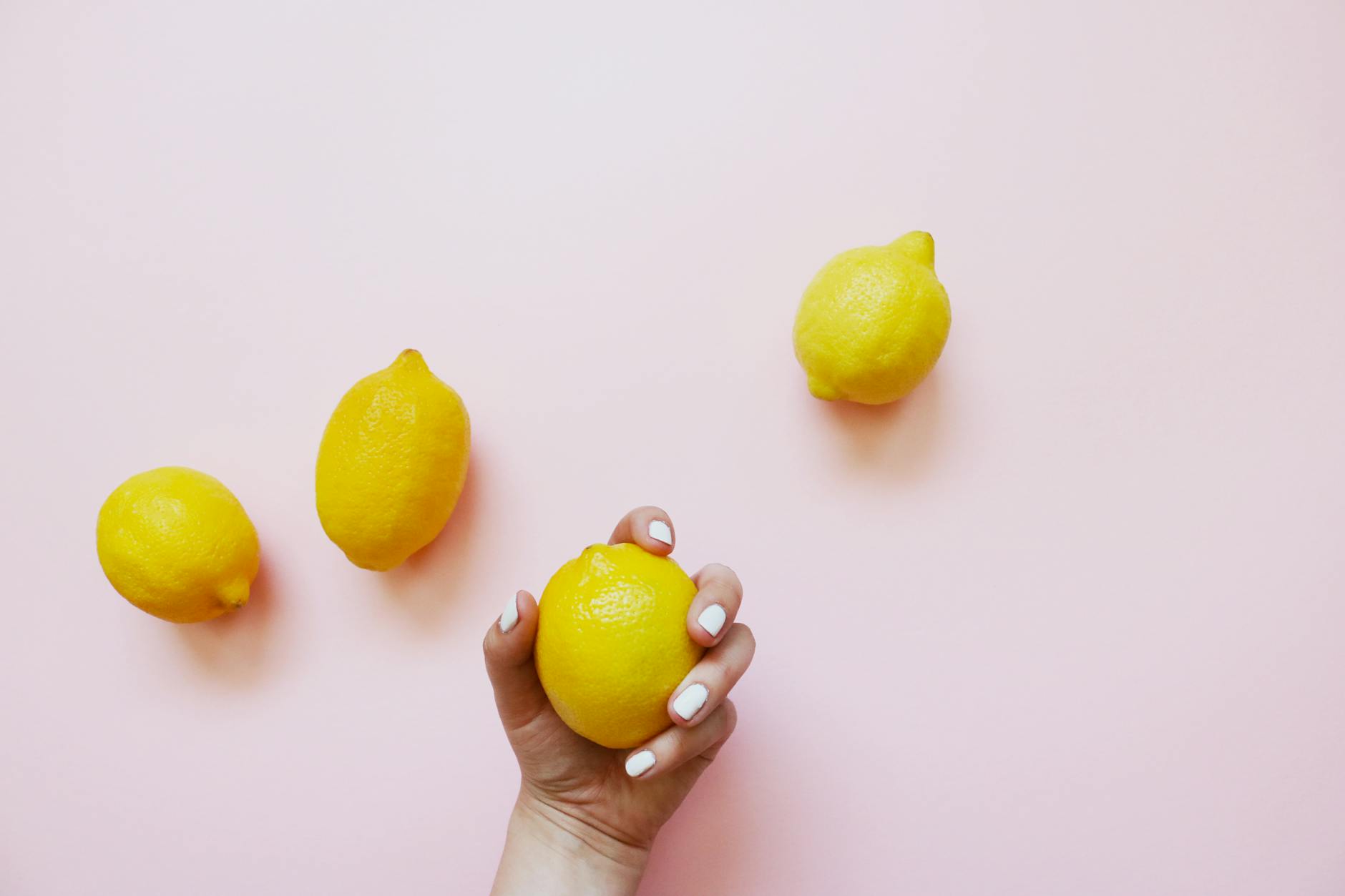 A hand holding a lemon on a soft pink background with fresh lemons nearby