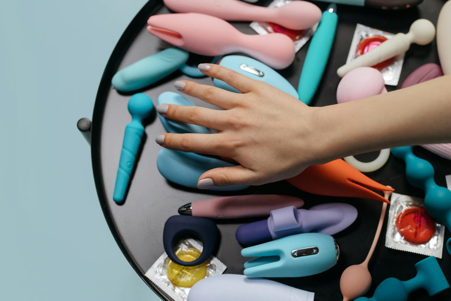Hand reaching toward a collection of colorful silicone vibrators and adult toys on a neutral surface