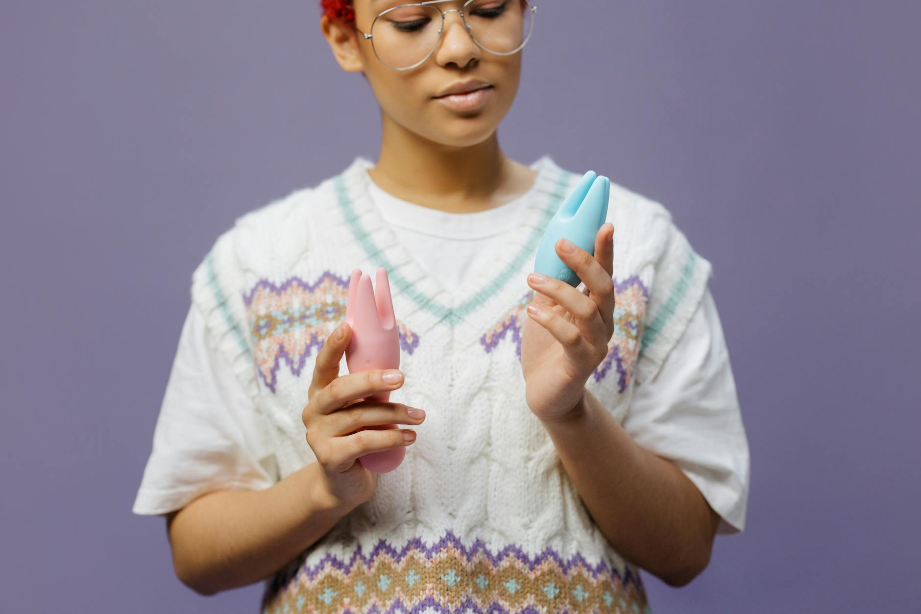 Woman in eyeglasses holding a blue and pink vibrator, looking thoughtful and intentional about her pleasure