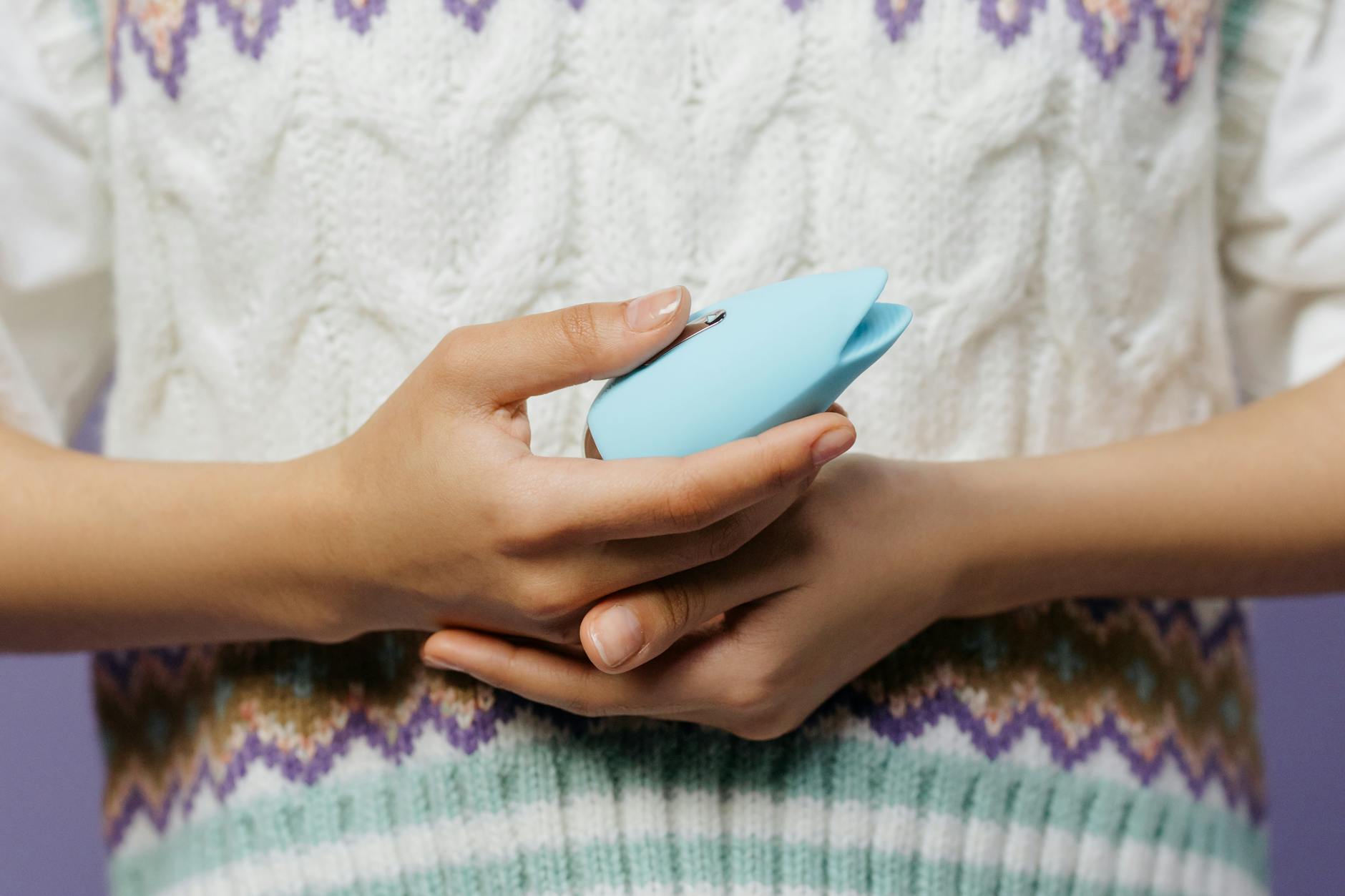 A hand with white nails holding a blue vibrator against a soft knitted sweater.